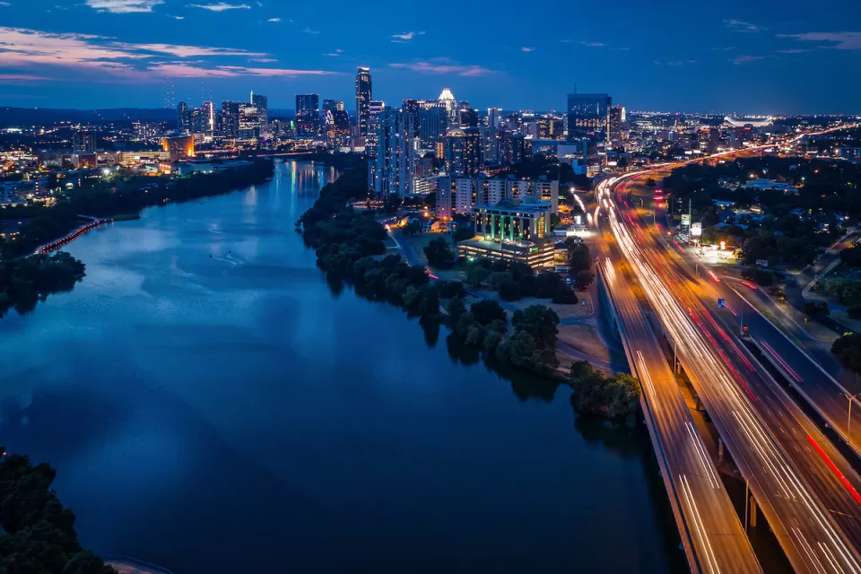 Aerial view of Interstate 35 with blurred traffic and Austin, Texas skyline in background with river in foreground at nighttime.